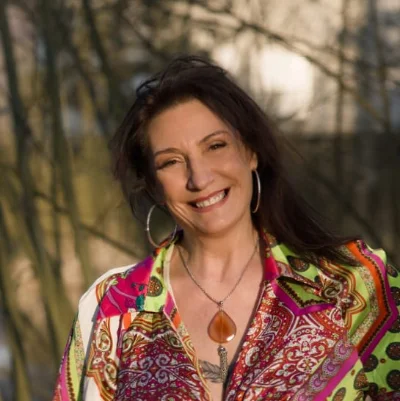 Femme souriante aux cheveux bruns, portant une chemise colorée et un pendentif, photographiée en extérieur